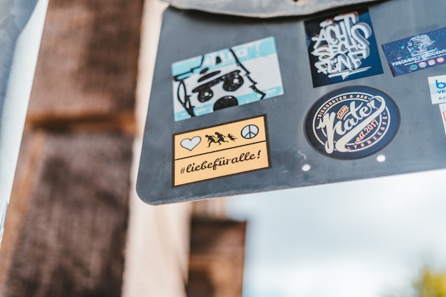A collection of colorful stickers is placed on a gray surface. Each sticker features different designs and logos, including a bear-like drawing, abstract text, and a peace symbol. One of the stickers displays the hashtag '#liebefüralle!' along with silhouettes of people running and a peace sign.