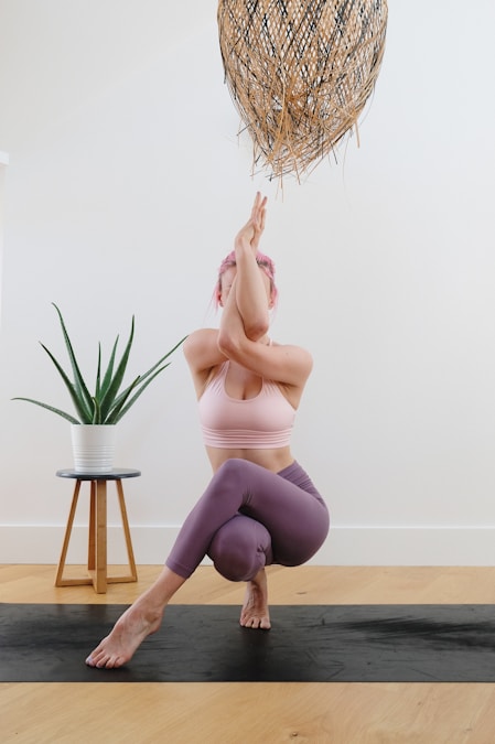 Woman with pink hair balancing in eagle pose on a black yoga mat in a bright studio