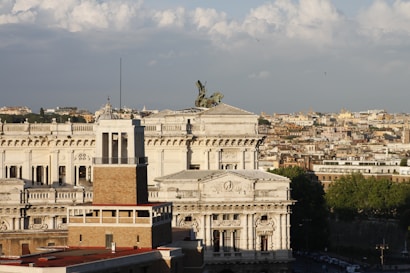 A panoramic view of a historic city featuring a grand neoclassical building with intricate architectural details and a bronze chariot statue on top. The foreground includes a modern brick tower with a flat roof, and the background showcases a sprawling urban landscape under a partly cloudy sky.