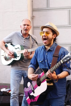 Two men are energetically playing guitars, one wearing a blue denim shirt and hat with yellow sunglasses, the other in a gray shirt. The man in front has a guitar with a unique design featuring a silhouette of a wolf and mountains.