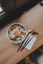 vegetable salad in white ceramic bowl beside stainless steel fork and knife on brown wooden table