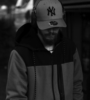 A person wearing a hooded jacket and a baseball cap with an 'NY' logo, listening to music through headphones. The background is blurred, suggesting an outdoor or urban setting. The image is in black and white, adding a dramatic feel.