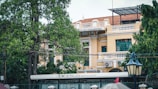 A two-story colonial-style building with a yellow facade and green window shutters. The building is partially obscured by lush green trees and has a large sign indicating it is a restaurant. A vintage street lamp is visible in the foreground.