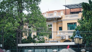 A two-story colonial-style building with a yellow facade and green window shutters. The building is partially obscured by lush green trees and has a large sign indicating it is a restaurant. A vintage street lamp is visible in the foreground.