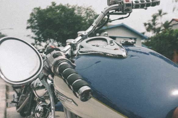 A close-up view of a motorcycle handlebar and fuel tank, showcasing shiny chrome elements and a classic design. The motorcycle is positioned outdoors, with blurred greenery and a house in the background, evoking a sense of adventure.