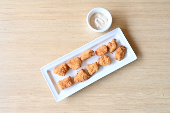 A white rectangular plate contains several pieces of golden-brown fried chicken nuggets arranged neatly. To the side, a small white bowl holds a light-colored, creamy dipping sauce. The background features a light wooden surface.