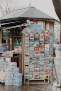 A small kiosk with a sloped roof features a display of colorful postcards and brochures, likely tourist-focused, with images of various landmarks and scenes. A stack of bottled water is neatly arranged on the ground beside the kiosk. Several printed materials are also visible, hinting at other items for sale within the kiosk.