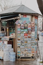 A small kiosk with a sloped roof features a display of colorful postcards and brochures, likely tourist-focused, with images of various landmarks and scenes. A stack of bottled water is neatly arranged on the ground beside the kiosk. Several printed materials are also visible, hinting at other items for sale within the kiosk.