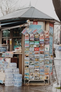 A small kiosk with a sloped roof features a display of colorful postcards and brochures, likely tourist-focused, with images of various landmarks and scenes. A stack of bottled water is neatly arranged on the ground beside the kiosk. Several printed materials are also visible, hinting at other items for sale within the kiosk.