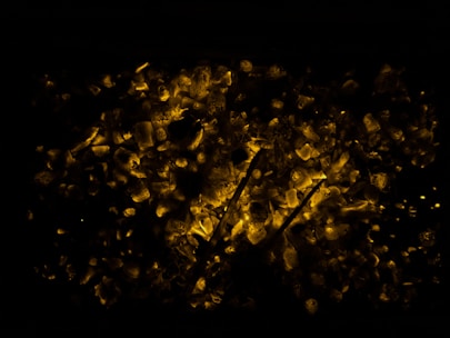 Glowing coals with a dark background create a warm, fiery effect. Randomly scattered, pieces of coal emit a bright orange-yellow hue, providing a contrast against the surrounding darkness.