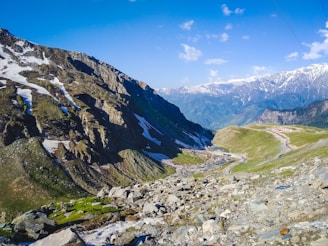 Rocky mountains with patches of snow under a clear blue sky, with a winding road and lush green valleys below.