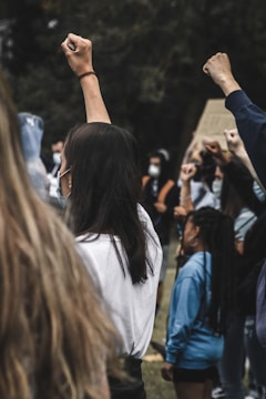 A group of people are raising their fists in a gesture of solidarity. Some are wearing masks and are standing outdoors, possibly in a park or public space. The mood is serious and focused, with individuals appearing engaged in a collective action.
