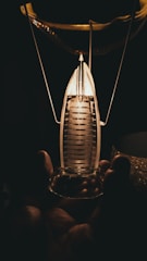 A hand holding a small, intricately designed glass object that resembles a miniature lantern or light bulb. The object emits a warm, glowing light that illuminates part of the surrounding darkness.
