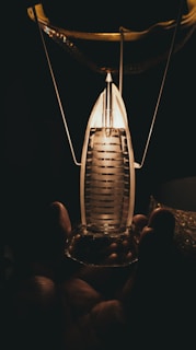 A hand holding a small, intricately designed glass object that resembles a miniature lantern or light bulb. The object emits a warm, glowing light that illuminates part of the surrounding darkness.