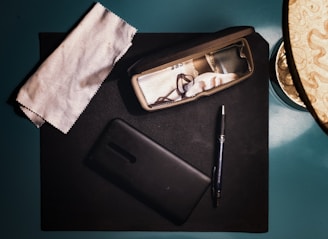 A flat lay of everyday objects including a cleaning cloth, a black case containing reading glasses, a smartphone, and a pen. The objects are arranged on a dark surface with ambient lighting casting soft shadows.