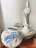 A white porcelain statue of a serene figure is placed on a textured stone platform. Next to it, there is a large decorative vase with blue and pink floral designs and butterfly motifs. The background is a neutral-colored wall, offering contrast to the intricately detailed objects.