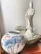 A white porcelain statue of a serene figure is placed on a textured stone platform. Next to it, there is a large decorative vase with blue and pink floral designs and butterfly motifs. The background is a neutral-colored wall, offering contrast to the intricately detailed objects.