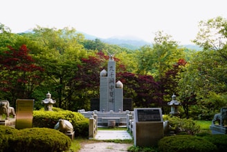 A stone monument stands surrounded by lush greenery and colorful trees in a serene, mountainous setting. The monument features inscribed text and is flanked by two decorative stone lanterns and several plaques. In the background, dense forest and distant mountains are visible under a hazy sky.