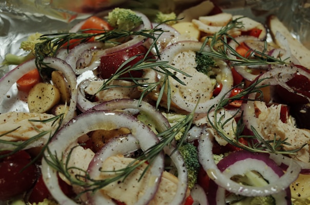 A close-up of a fresh and colorful salad with various vegetables and herbs. The salad includes sliced onions, broccoli florets, cherry tomatoes, bell peppers, and what appears to be grilled chicken pieces. The ingredients are garnished with sprigs of rosemary and sprinkled with ground black pepper.