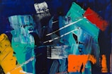 A vibrant abstract painting with bold brushstrokes and contrasting colors, including teal, blue, orange, and touches of red and yellow. The composition features dynamic, sweeping lines and textured surfaces that create a sense of movement and energy.
