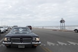 A classic car with a Texas license plate is prominently driving down a coastal road. The scene includes multiple other vehicles, mostly modern, driving in the same direction. To the right, there is a view of the ocean with waves and a cloudy sky above. A large signpost is visible by the roadside.