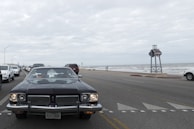 A classic car with a Texas license plate is prominently driving down a coastal road. The scene includes multiple other vehicles, mostly modern, driving in the same direction. To the right, there is a view of the ocean with waves and a cloudy sky above. A large signpost is visible by the roadside.