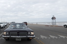 A classic car with a Texas license plate is prominently driving down a coastal road. The scene includes multiple other vehicles, mostly modern, driving in the same direction. To the right, there is a view of the ocean with waves and a cloudy sky above. A large signpost is visible by the roadside.