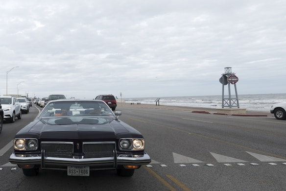 A classic car with a Texas license plate is prominently driving down a coastal road. The scene includes multiple other vehicles, mostly modern, driving in the same direction. To the right, there is a view of the ocean with waves and a cloudy sky above. A large signpost is visible by the roadside.