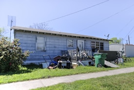 A small, single-story house with blue siding and a flat roof is shown. The front yard features a green trash bin, a small lawn mower, and various scattered items, including a bicycle. An old satellite dish is mounted on the roof, and there are electrical lines visible in the background. The grass is lush and green, and there's a concrete pathway leading up to the house.