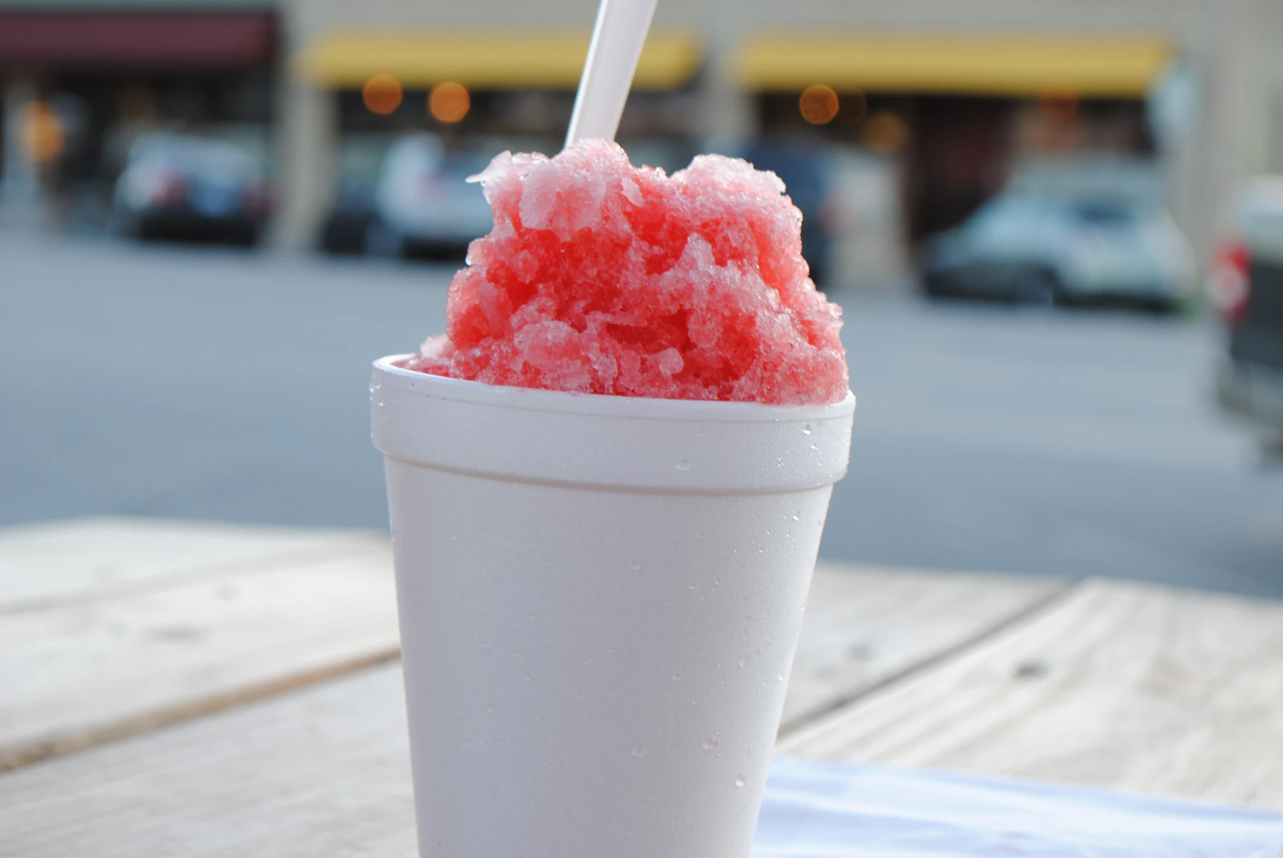A coarse red snow cone heaped in a foam cup with a spoon straw, on a picnic table by a street