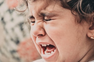 A close-up of a young child with curly hair, expressing a strong emotion. The child’s eyes are tightly closed, and the mouth is open wide, suggesting crying or shouting. The skin tone is soft, and the earring adds a subtle touch to the image.