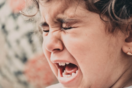 A close-up of a young child with curly hair, expressing a strong emotion. The child&rsquo;s eyes are tightly closed, and the mouth is open wide, suggesting crying or shouting. The skin tone is soft, and the earring adds a subtle touch to the image.