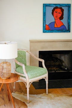 A modern interior space features a wooden chair with a patterned green cushion next to a small round wooden table. On the table is a lamp with a woven rattan base and a white lampshade. There is a colorful painting hanging above a fireplace with a light mantel. The floor has a light wood finish and a light-colored animal hide rug partially visible.