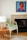 A modern interior space features a wooden chair with a patterned green cushion next to a small round wooden table. On the table is a lamp with a woven rattan base and a white lampshade. There is a colorful painting hanging above a fireplace with a light mantel. The floor has a light wood finish and a light-colored animal hide rug partially visible.