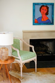 A modern interior space features a wooden chair with a patterned green cushion next to a small round wooden table. On the table is a lamp with a woven rattan base and a white lampshade. There is a colorful painting hanging above a fireplace with a light mantel. The floor has a light wood finish and a light-colored animal hide rug partially visible.