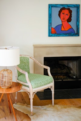 A modern interior space features a wooden chair with a patterned green cushion next to a small round wooden table. On the table is a lamp with a woven rattan base and a white lampshade. There is a colorful painting hanging above a fireplace with a light mantel. The floor has a light wood finish and a light-colored animal hide rug partially visible.