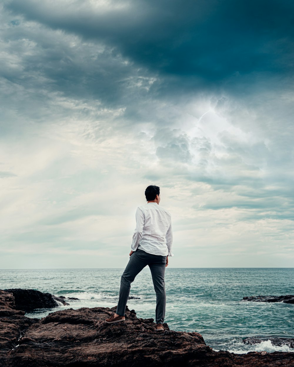 man in white dress shirt standing on brown rock near body of water during daytime