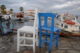 Two wooden chairs, one painted white and the other blue, are placed on a stone-paved waterfront. Several boats, including fishing and leisure vessels, are moored in the calm water of the harbor. In the background, palm trees and buildings with red and white colors add to the coastal atmosphere under a partly cloudy sky.