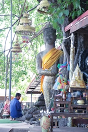 A large statue of Buddha wrapped in a golden cloth stands near a tree with green leaves. Various smaller religious items, including a small Buddha statue and other decorative objects, are placed around the statue. A man is kneeling in prayer near the statue, wearing a blue shirt. Hanging ornate decorations are visible from the branches of the tree.