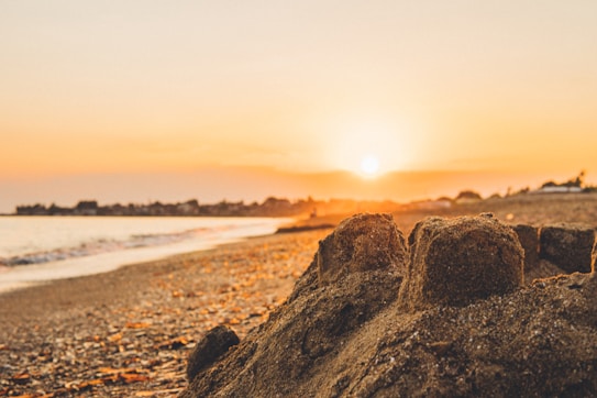 A serene beach scene at sunset with a golden glow illuminating the sky and the sandy shore. The sun is low on the horizon, casting warm hues across the scene. In the foreground, a partially crumbled sandcastle is visible, adding a playful element to the tranquil setting.