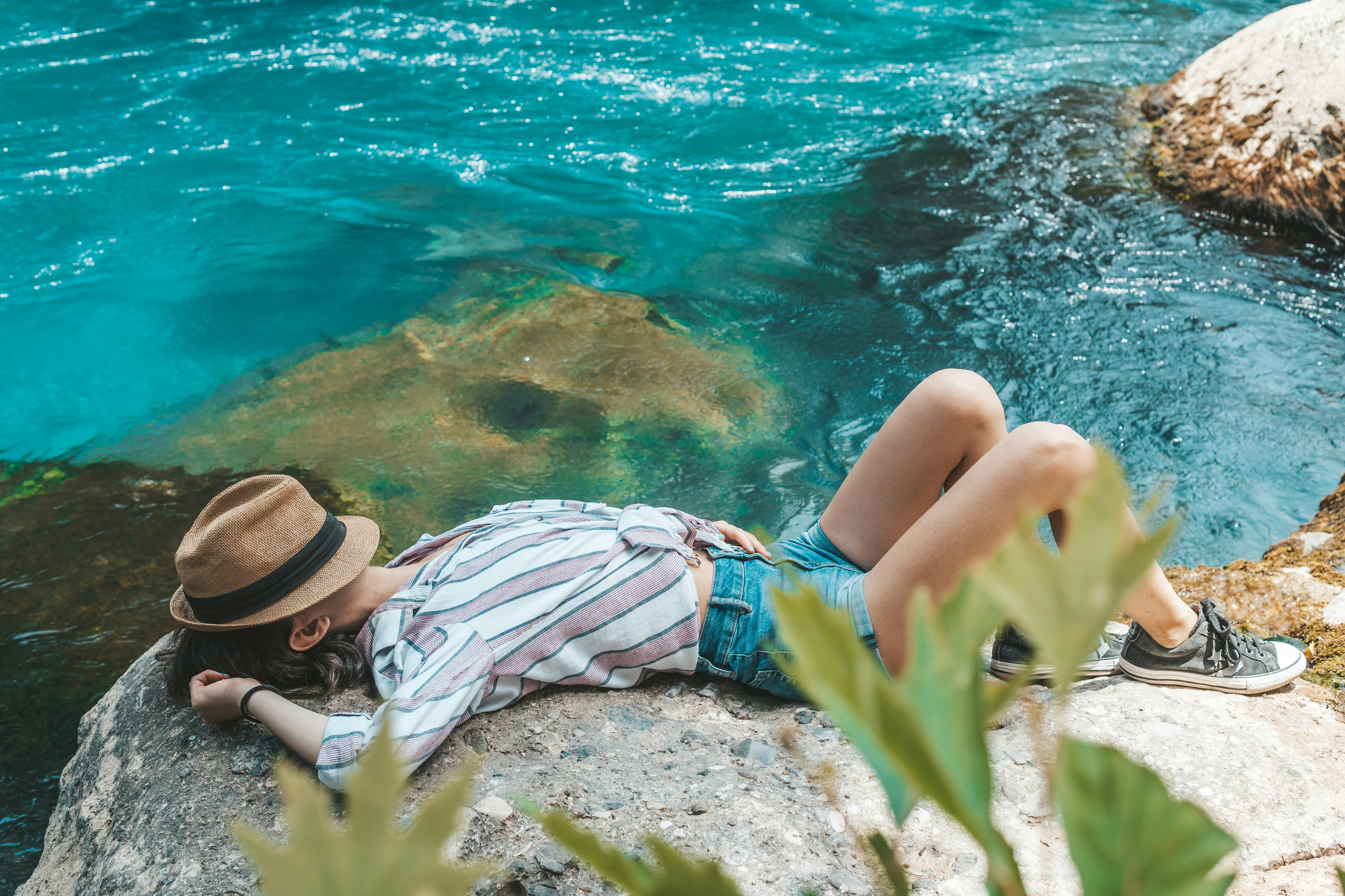 woman in blue denim daisy dukes and brown hat sitting on rock near body of water