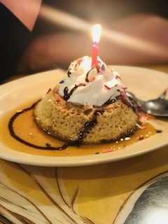 A dessert topped with whipped cream and colorful sprinkles, featuring a lit candle at the center. The dessert is served on a plate with chocolate drizzle.
