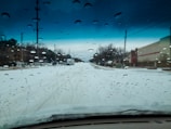 Raindrops are scattered across a car windshield, while a snow-covered road stretches ahead. Telephone poles line the street, and a large commercial building is visible on the right. Overcast skies add an ominous tone, with a gradient of dark clouds on the horizon.