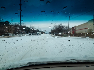 Raindrops are scattered across a car windshield, while a snow-covered road stretches ahead. Telephone poles line the street, and a large commercial building is visible on the right. Overcast skies add an ominous tone, with a gradient of dark clouds on the horizon.