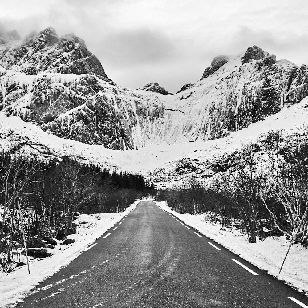 grayscale photo of road between snow covered trees and mountains
