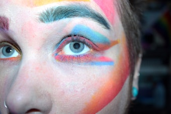 A close-up of a person's face with vibrant, multicolored makeup applied around the eye, including shades of blue, red, orange, and yellow. The eyebrow is dark, and the eye itself is prominently visible with a piercing gaze.