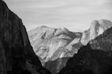 A black and white photograph of a mountainous landscape, featuring dramatic contrasts between light and shadow on the rugged cliffs and slopes. The central mountain has a distinct, smooth face that stands out against the textured foreground and background peaks.