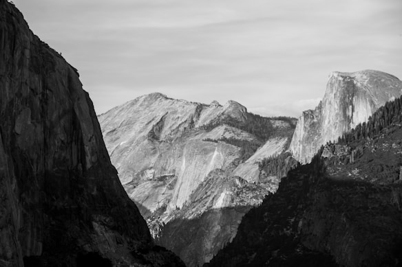 A black and white photograph of a mountainous landscape, featuring dramatic contrasts between light and shadow on the rugged cliffs and slopes. The central mountain has a distinct, smooth face that stands out against the textured foreground and background peaks.