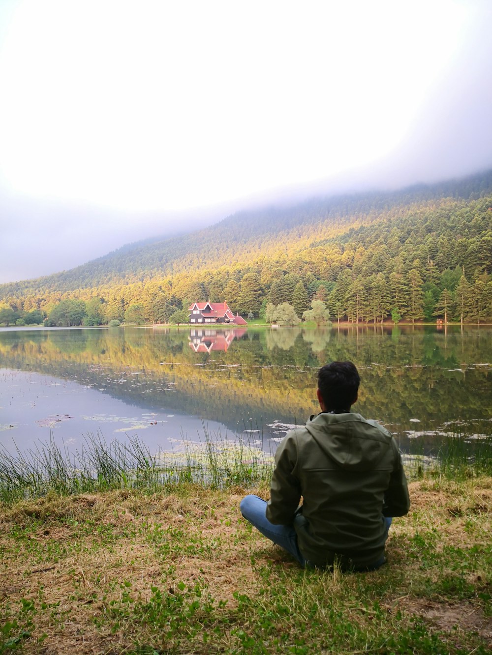 uomo in giacca grigia che si siede sull'erba verde vicino al lago durante il giorno