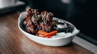 A white dish containing pieces of crispy, glazed chicken wings garnished with green herbs. Accompanied by carrot and celery sticks, along with a small black container of dipping sauce. The wooden table adds a rustic feel.